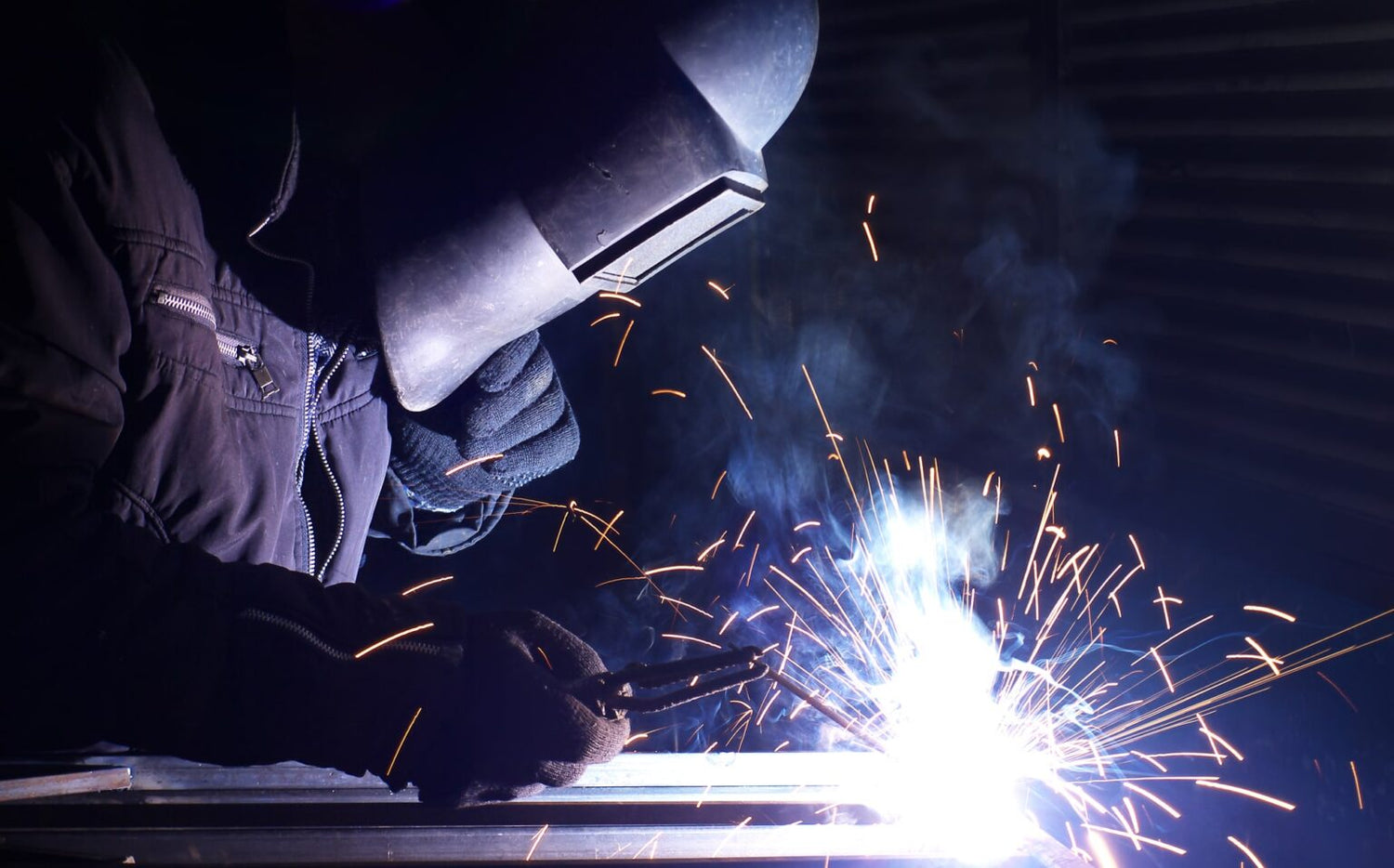 Welder wearing protective gear working with bright sparks in a dark workshop